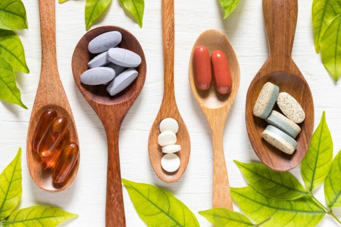 Five wooden spoons holding various supplements—soft gels, gray tablets, white pills, red capsules, and speckled tablets.