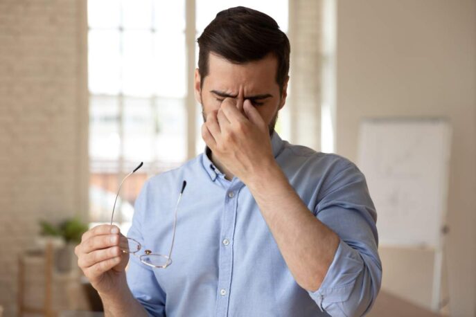 Man holding glasses and pinching the bridge of his nose with eyes closed, appearing tired or eye-strained in a bright room.