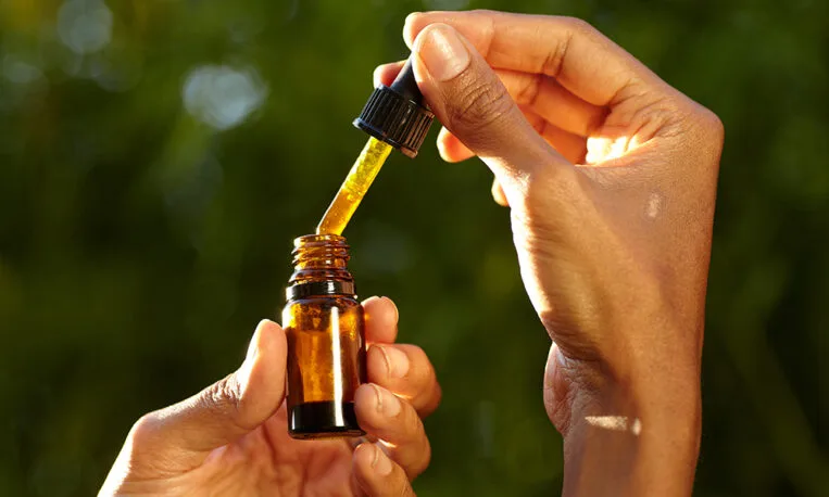 Close-up of hands holding an amber glass dropper bottle, lifting a dropper of golden oil against a blurred green background.